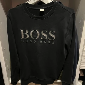 Svart sweatshirt från Hugo Boss - Svart sweatshirt från Hugo Boss med stor BOSS-logga i grått framtill. Tröjan har rund halsringning och långa ärmar. Tillverkad i mjukt bomullsmaterial som är skönt mot huden. Perfekt för en stilren och avslappnad look.