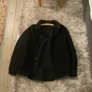 Svart overshirt från Uniqlo - Stilren svart overshirt från Uniqlo i storlek XL. Skjortan har klassisk krage, knäppning framtill och långa ärmar. Perfekt att slänga över en t-shirt för en clean look. Materialet känns mjukt och lätt, troligtvis bomull eller polyester.