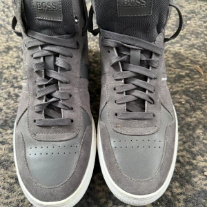 Grå sneakers från Hugo Boss, strl 40 - Snygga grå sneakers från Hugo Boss med vita sulor och diskreta logodetaljer. Skorna har ovandel i mocka och skinn, klassisk snörning och rund tå. Perfekta för dig som gillar stilrena och sportiga vibes.