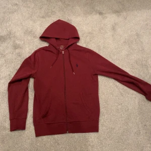 Vinröd hoodie från Polo Ralph Lauren - Snygg vinröd hoodie från Polo Ralph Lauren med dragkedja och huva. Tröjan har långa ärmar, snörning i huvan och klassisk blå logga broderad på bröstet. Perfekt för en chill och stilren look.