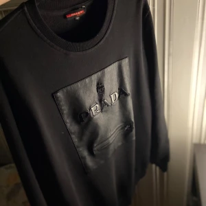 Svart Prada sweatshirt med skinnficka - Svart sweatshirt från Prada med stor skinnficka framtill, broderad logga och dragkedjedetalj. Tröjan har rund halsringning och långa ärmar. Snygg och stilren design som sticker ut med exklusiva materialval.  Det står att det är en storlek 2XL men passar som medium kanske även L, Äkta det vet jag inte! Kom med ett pris så är den din🤩