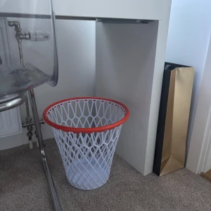 Basketkorg papperskorg  - Säljer en vit papperskorg med röd/orange kant, formad i ett stilrent nätmönster som ger en luftig och modern känsla. Designen påminner om en basketkorg och gör den både praktisk och dekorativ.  Den är lätt, rymlig och enkel att placera under skrivbordet eller i barnrummet, men fungerar lika bra i hemmakontoret eller vardagsrummet. Väl använd men i mycket gott skick. Skriv gärna vid frågor, pris kan diskuteras😊😊