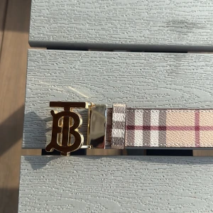 Burberry rutigt skärp med guldspänne - Snyggt Burberry-bälte i klassiskt rutigt mönster med beige, bruna, vita och röda toner. Skärpet har ett exklusivt guldigt spänne med Burberrys ikoniska monogram. Tillverkat i skinn med lyxig känsla och detaljerad finish.