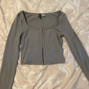 Grå ribbad croppad topp med hyskor från hm - Snygg grå croppad topp med långa ärmar och ribbad struktur. Toppen har en djup, rundad urringning och stängs framtill med hyskor för en cool detalj. Perfekt att styla med jeans eller kjol för en trendig look.  Priset går att diskutera 