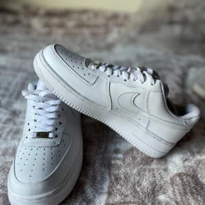 Nike Air Force 1 vita sneakers - Klassiska Nike Air Force 1 i helvitt skinn med perforerad tå, vit sula och snörning. Snygg och tidlös modell med låg profil och rund tå. Perfekta för dig som vill ha en clean och stilren look. Kommer med originalpåse från Nike.