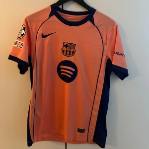 Barcelona Lamine Yamal Nike fotbollströja - Snygg FC Barcelona fotbollströja i orange med mörkblå detaljer, Nike-logga och klubbmärke på bröstet. Tryck med Lamine Yamal och nummer 19 på ryggen. Korta ärmar, officiella sponsorer och Champions League-märke på ärmen. Tillverkad i lätt och ventilerande material. Replika