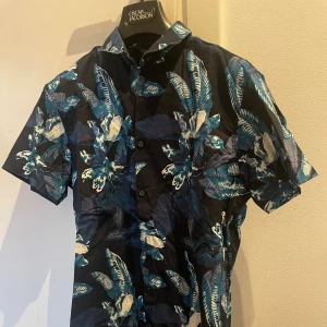 Blommig kortärmad skjorta Selected Homme  - Snygg kortärmad skjorta från Selected Homme  med blått och vitt blommönster på mörk botten. Skjortan har klassisk krage och knäppning framtill. Perfekt för dig som vill sticka ut med en färgstark och trendig look.