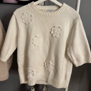 Vit stickad tröja med blommor Holly & Whyte - Superfin vit stickad tröja från Holly & Whyte med korta ärmar och stora blommiga applikationer framtill. Tröjan har ribbade muddar vid hals, ärmslut och nederkant. Strl är L men upplevs/sitter som en XS/S.