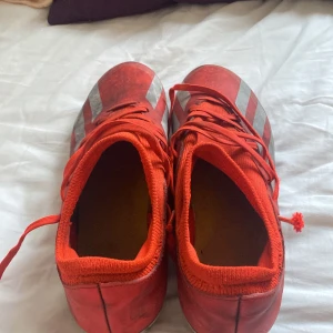 Röda Adidas fotbollsskor strl 42 - Snygga röda Adidas fotbollsskor med silvriga ränder på sidorna och snörning framtill. Skorna har en strumpliknande mudd och platt sula med dobbar för bra grepp på planen. Perfekta för dig som vill sticka ut på matchen!
