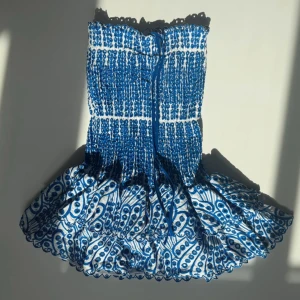 Blåvit kortklänning med volang - Supersöt blåvit kortklänning med smockad överdel och volangig kjol. Klänningen har ett unikt mönster med blå broderade detaljer och knytband framtill. Perfekt för varma dagar och ger en riktigt fräsch sommarkänsla. Fodrad insida för extra komfort. Storlek XL men de är nog mer onesize💙