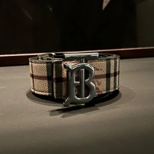 Burberry bälte med rutig design - Snyggt Burberry bälte med klassiskt rutig mönster i beige, svart och rött. Bältet har en stor silvrig metallspänne med Burberrys ikoniska B-logga. Utsidan är i mönstrat tyg och insidan i svart skinn. Perfekt accessoar för att lyfta din outfit.                                                   Använt en gång ! 110cm
