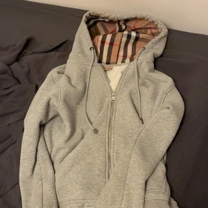 Grå hoodie från Burberry med dragkedja - Säljer en grå hoodie från Burberry i bomull med klassiskt rutigt foder i huvan. Tröjan har dragkedja framtill, snörning i huvan och broderad logga på bröstet. Perfekt för dig som gillar stilrena och exklusiva plagg.