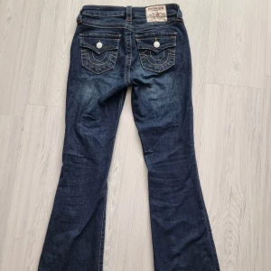 True Religion bootcut jeans blå - Snygga blå bootcut jeans från True Religion, står att de är 25(XXS) men passar mig som är XS/S. Jeansen har klassiska kontrastsömmar, coola ficklock med silverfärgade knappar och tydlig logga bak. Materialet är jeans med en mörk tvätt och orange detaljer vid fickorna.