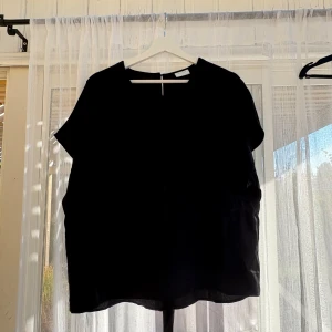 Svart blus med knyt från Vila - Svart blus från Vila med korta ärmar och rund halsringning. Baktill finns en öppning med knytband i midjan som ger en snygg detalj. Tunt, lätt tyg som känns luftigt och passar perfekt till många olika looks.