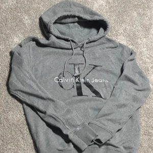Grå hoodie Calvin Klein Jeans - Snygg grå hoodie från Calvin Klein Jeans med stor CK-logga framtill. Tröjan har huva med snörning och långa ärmar med ribbade muddar. Mjuk bomullsblandning som är perfekt för chill dagar. Enkel och stilren design som funkar till allt.