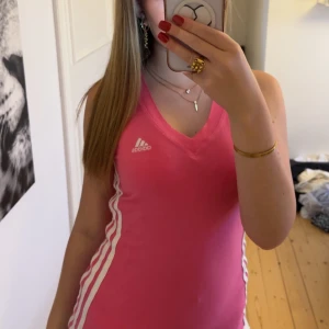 Rosa Adidas topp - Sportig rosa linnetopp från Adidas med v-ringning och vita ränder längs sidorna. Perfekt för träning eller tennis, tillverkad i mjuk bomullsmix. Klassisk Adidas-logga på bröstet och ärmlös design för maximal rörelsefrihet.