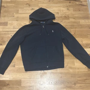 Blå hoodie från Polo Ralph Lauren - Svart hoodie från Polo Ralph Lauren med klassisk broderad logga på bröstet. Tröjan har huva med snörning, dragkedja framtill och två fickor. Perfekt för en chill och stilren look.