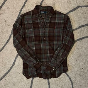 Rutig skjorta från Polo Ralph Lauren - Snygg rutig skjorta från Polo Ralph Lauren i brunt, grönt och rött. Klassisk button-down krage, långa ärmar och knäppning framtill. Skjortan har en broderad logga på bröstet och är tillverkad i mjuk bomull. Perfekt för dig som gillar en avslappnad men stilren look.