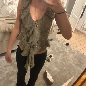 Olivgrön volangblus med knyt - Supersöt olivgrön blus med volanger och  v-ringning. Toppen har en crinklad struktur och knyt framtill. Perfekt för dig som gillar detaljer. Ärmlös och croppad modell som passar till både jeans och kjol.