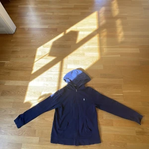Marinblå hoodie från Polo Ralph Lauren - Snygg marinblå hoodie från Polo Ralph Lauren med dragkedja framtill och klassisk huva. Tröjan har långa ärmar, två fickor och den ikoniska lilla loggan broderad på bröstet. Perfekt för dig som gillar stilren och sportig look.