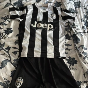 Juventus Tevez 10 fotbollströja set - Juventus matchtröja med Tevez 10 på ryggen och klassiska svarta och vita ränder. Jeep-logga på bröstet och klubbmärke både på tröjan och shortsen. Kortärmad modell i lätt och ventilerande material, perfekt för fotbollsträning eller match. För barn