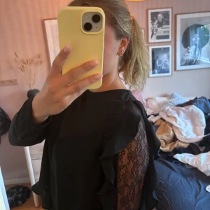 Svart blus med spets och volang - Säljer en svart blus med långa ärmar, snygga volangdetaljer och inslag av spets längs ärmarna. Blusen har rund halsringning och knäppning vid ärmslut. Perfekt för dig som gillar en stilren look med lite extra detaljer.
