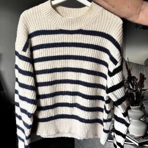 Randig stickad tröja i vitt och marinblått - Säljer en ribbstickad tröja med breda marinblå ränder på vit botten. Tröjan har rund hals och långa ärmar, perfekt för dig som gillar en chill och klassisk look. Materialet är mjukt och stickat, vilket gör den extra skön att bära under kyliga dagar.