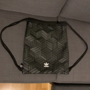 Svart gympåse från Adidas med mönster - Snygg svart gympåse från Adidas med geometriskt mönster på framsidan och klassisk logga. Väskan har snörstängning och en praktisk dragkedjeficka på sidan. Tillverkad i slitstark polyester, perfekt för träning eller skola.