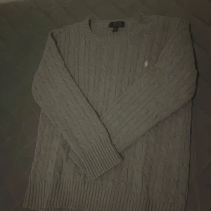 Grå kabelstickad tröja Polo Ralph Lauren - Kabelstickad grå tröja från Polo Ralph Lauren med klassisk rund hals och broderad logga på bröstet. Tröjan har långa ärmar och breda ribbade muddar vid ärmslut och nederkant. Perfekt för lager-på-lager och chill höst- och vinterstil.