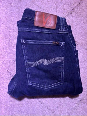 Mörkblå Nudie Jeans 30/32 - Snygga mörkblå jeans från Nudie Jeans i klassisk rak modell. Jeansen har kontrastsömmar, fem fickor och Nudie-logga på bakfickan samt läderpatch i midjan. Materialet är slitstark bomull och passformen är normal med straight legs. Passar 160-170cm