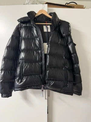 Svart Moncler pufferjacka med huva Storlek L - Säljer en svart pufferjacka från Moncler med glansig finish och huva. Jackan har dragkedja framtill, två sidofickor med dragkedja och en ficka med Moncler-logga på ärmen. Perfekt för kalla dagar och har en riktigt schysst streetstil. aldrig använt