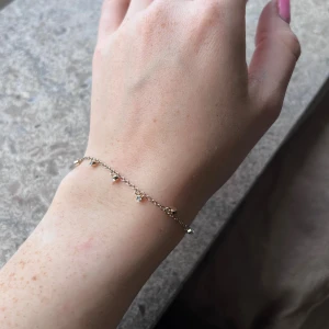 Pilgrim armband - Superfint och nätt armband från Pilgrim i guldtonad metall med små gnistrande stenar som hänger längs kedjan. Perfekt för dig som gillar minimalistiska smycken med lite extra bling. Justerbar längd med Pilgrim-tag vid låset. Knappt använt. En liten sten har ramlat av. Syns bäst på 3:e bilden där avståndet mellan stenarna är lite längre på ett ställe.