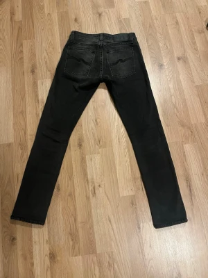 Svarta jeans från Nudie Jeans Co - Snygga svarta jeans från Nudie Jeans Co med klassisk femficksdesign och svarta broderade detaljer på bakfickorna. Jeansen har en rak passform och är tillverkade i mjukt bomullsmaterial med en mörk tvätt. Perfekta för dig som gillar stilrena och tidlösa jeans.