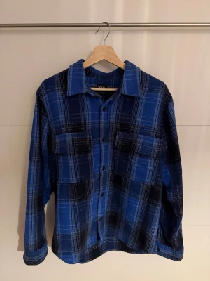 Blå rutig skjorta från Zara - Säljer en blå och svart rutig skjorta från Zara med klassisk krage och knappar framtill. Skjortan har två bröstfickor och långa ärmar. Perfekt för dig som gillar en chill och stilren look. Materialet är mjukt och känns skönt mot huden.