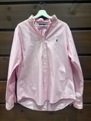 Rosa randig skjorta från Polo Ralph Lauren - Snygg rosa skjorta med vita ränder från Polo Ralph Lauren. Klassisk button-down krage, långärmad och med broderad marinblå logga på bröstet. Tillverkad i mjuk bomull och har knappar framtill. Perfekt för dig som gillar stilrena och fräscha plagg. STORLEK: M/L