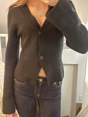 Svart kofta med v-ringning och knappar - Snygg svart kofta med v-ringad hals och stora knappar framtill. Koftan har långa ärmar med breda muddar och är tillverkad i ett mjukt material som känns skönt mot huden. Perfekt att slänga på sig över en topp för en chill look.