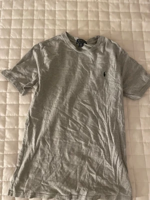 Grå t-shirt från Polo Ralph Lauren - Säljer en enkel och stilren grå t-shirt från Polo Ralph Lauren med klassisk rund hals och korta ärmar. T-shirten har en liten broderad logga på bröstet och är gjord i mjuk bomull som är skön mot huden. Perfekt för en chill och avslappnad look.