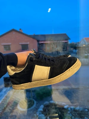 Valentino Garavani svarta sneakers - Säljer ett par snygga Valentino Garavani sneakers i svart och vitt. Skorna har en klassisk låg profil, rund tå och platt sula. Ovandelen är i mocka och skinn med svarta snören och vit panel på sidan. Märkesdetaljer på plösen och yttersulan.