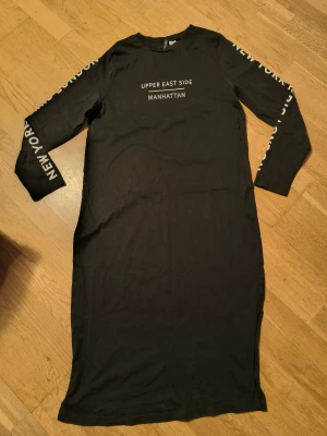 Svart långklänning med tryck - Svart långklänning med vita tryck längs ärmarna, Klänningen har lång ärm och är gjord i mjuk bomull, perfekt för en chill och urban look.