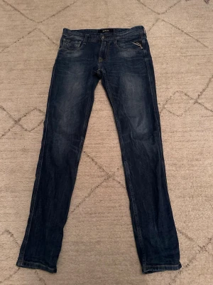 Replay anbass jeans - Snygga blå jeans från Replay med klassisk femficksdesign och diskreta slitningar. Modellen är slim fit med raka ben och normal midja. Jeansen har detaljer som Replay-märke på bakfickan och läderpatch i midjan. Perfekta för dig som gillar stilrena och tidlösa jeans.