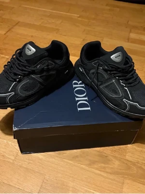 Svarta Dior sneakers med silverdetaljer - Säljer ett par svarta Dior sneakers med snygga silverdetaljer och logga på plösen. Skorna har rund tå, snörning och en platt sula. Materialet är en mix av mesh och syntet, vilket ger dem en modern och sportig vibe. Perfekta för dig som gillar exklusiva märken och streetstyle.