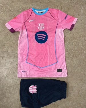 FC Barcelona fotbollströja och shorts - Säljer ett FC Barcelona matchställ med rosa tröja och mörkblå shorts. Tröjan har blå detaljer, klubbmärke, sponsortryck och nummer 10 med Lamine Yamal på ryggen. Tillverkad i polyester med Dri-Fit-teknik för bästa komfort på planen.