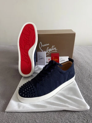 Christian Louboutin skor  - Säljer ett par mörkblå sneakers från Christian Louboutin med vit sula och ikonisk röd undersida. Skorna har snörning och är tillverkade i mocka med coola svarta nitar på tån. Insidan är beige och mjuk, perfekt för dig som gillar lyxiga detaljer.