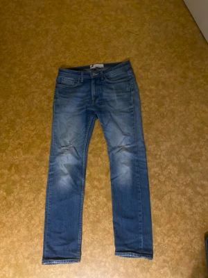 Blå jeans med slitningar - Säljer ett par blå jeans med snygga slitningar vid knäna och klassisk femficksdesign. Jeansen har en rak passform och är tillverkade i mjukt denimtyg. Färgen är mellanblå med lite ljusare tvättade partier för en cool look.