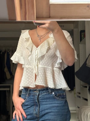 Zara blus - INTRESSEKOLL! Supersöt slutsåld vit blus från Zara i storlek XS med volangdetaljer längs halsringningen och ärmarna. Blusen har v-ringning, knappar framtill och är lätt transparent med korta ärmar. Perfekt för dig som gillar romantisk och feminin stil🥰