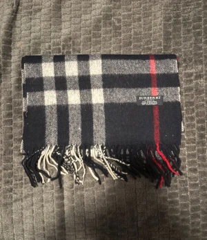 Rutig kashmirhalsdukfrån Burberry - Säljer en klassisk rutig halsduk från Burberry i 100% kashmir. Västen har en snygg kombination av mörkblå, grå, vit och röd färg med fransar längst ner. Perfekt för dig som vill ha en tidlös och lyxig look med brittisk touch.  Inköpt i bulk från en leverantör och har genomgått deras äkthetskontroll, vilket gör den till en pålitlig produkt. Jag kan dock inte personligen garantera 100 % autenticitet.