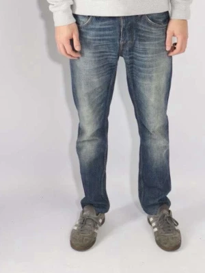Tiger of Swesen blå jeans straight fit - Säljer ett par klassiska Tiger of Sweden jeans i blå tvätt med snygga slitningar framtill. Modellen har rak passform och fem fickor, samt knappgylf. Jeansen är tillverkade i robust denim och har en tidlös look som funkar till många stilar.