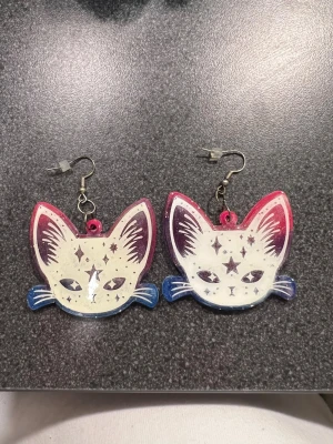 Färgglada kattörhängen  - Unika örhängen i form av kattansikten med stjärnmönster. Framsidan är vit med detaljer i mörklila, rosa och blå, medan baksidan har en gradient i rosa, lila och blå. med metallkrokar. Perfekt för dig som gillar statement-smycken och djurmotiv.