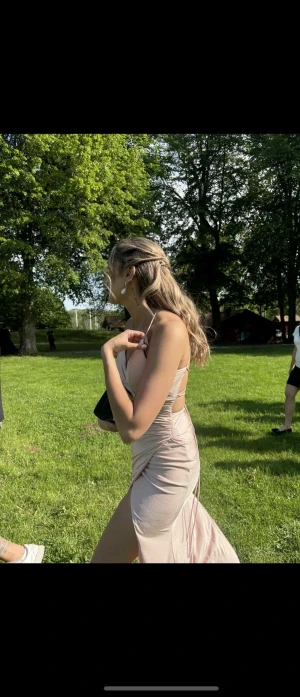 Ljusrosa balklänning - Säljer en elegant ljusrosa långklänning med omlott-design och djup slits framtill. Klänningen har smala axelband, öppen rygg med snörning och ett figurnära fall. Materialet är mjukt och har en lätt glansig finish som ger en lyxig känsla.