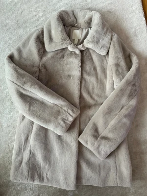Beige pälsjacka från H&M - Säljer en fluffig och mjuk beige pälsjacka från H&M med klassisk krage och raka långa ärmar. Jackan har en stilren look och är perfekt för dig som vill ha en cozy vibe under kyliga dagar. Fodrad insida för extra komfort.